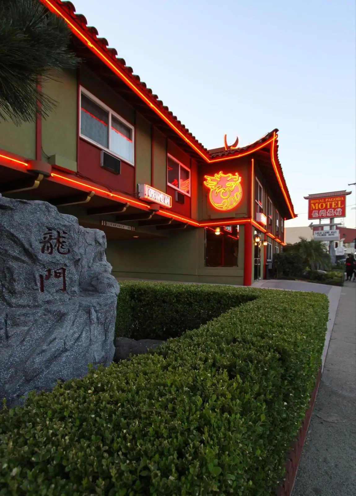 A green and red building with a pagoda-style roof and neon lights, a large rock with Chinese characters, and well-manicured hedges.