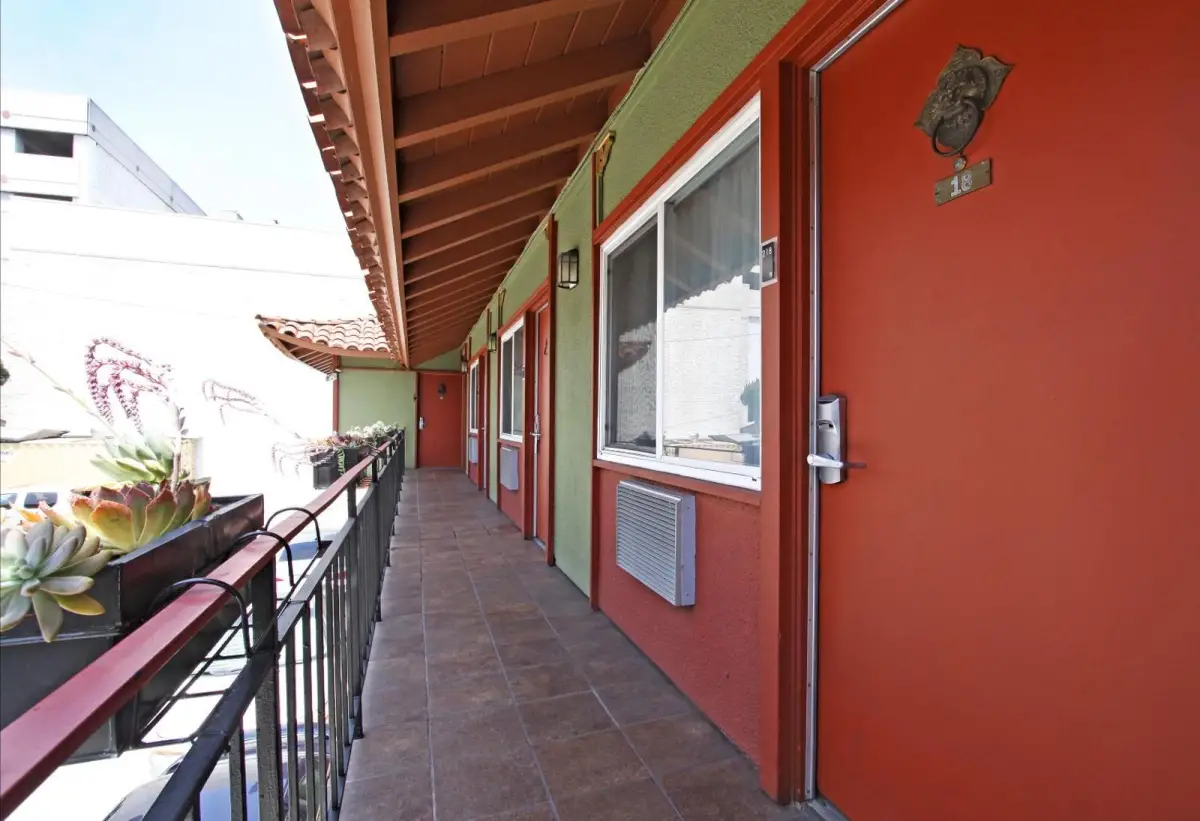 A long, narrow balcony with a tiled floor and red doors leads to a row of rooms with green walls and brown ceilings.
